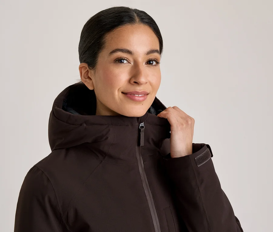 Femme souriante portant une veste à capuche marron foncé, tenant le col avec sa main.