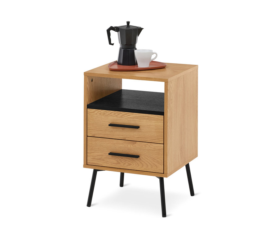 Table de chevet en bois clair avec deux tiroirs et une étagère ouverte, dotée de pieds et de poignées de tiroir noirs. Une cafetière moka noire et une tasse blanche sont posées sur un plateau brun-rouge sur le dessus.