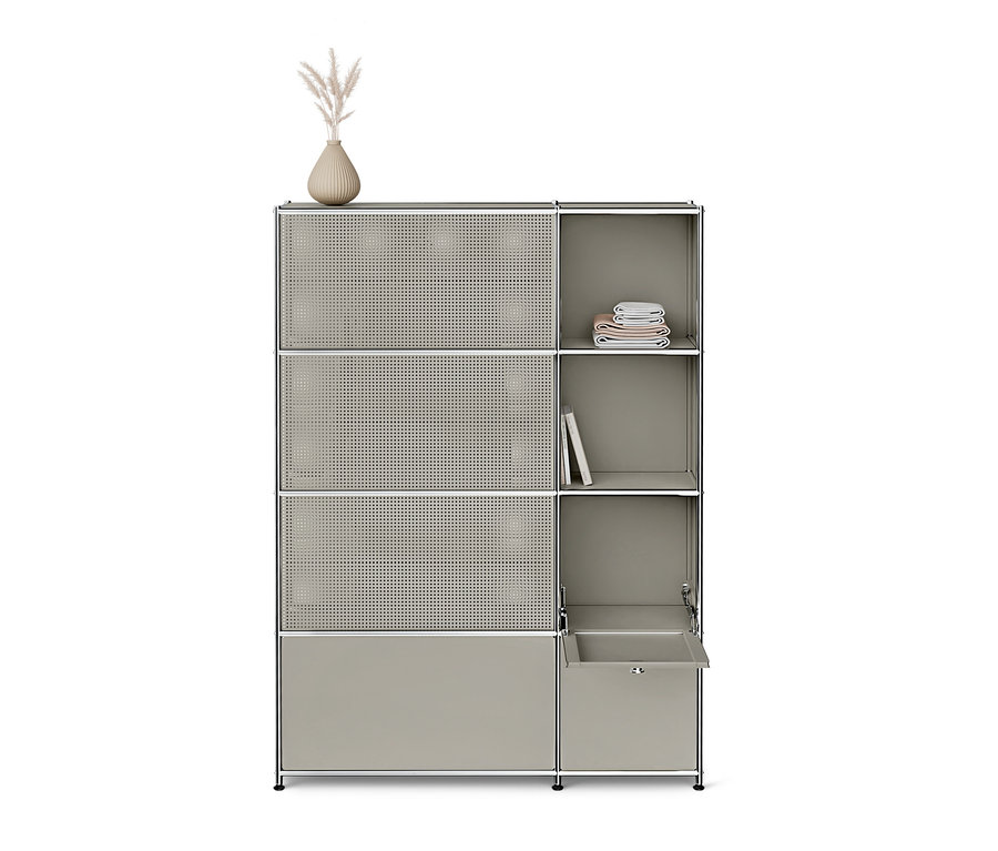 Une haute étagère modulaire grise avec un cadre argenté. À gauche, trois portes perforées et une porte pleine en bas. À droite, deux étagères ouvertes avec des serviettes pliées et un livre, et un tiroir partiellement ouvert en bas. Un vase avec de l'herbe séchée est posé dessus.