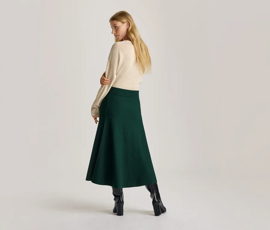 Femme vue de dos, regardant par-dessus son épaule, portant un pull crème, une jupe trapèze vert foncé et des bottes noires à talons.