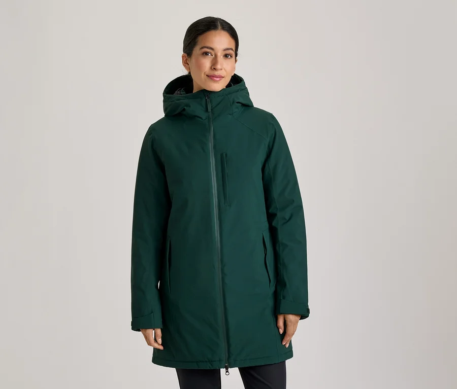 Femme debout, portant un manteau à capuche vert foncé zippé, les mains dans les poches, regardant la caméra.
