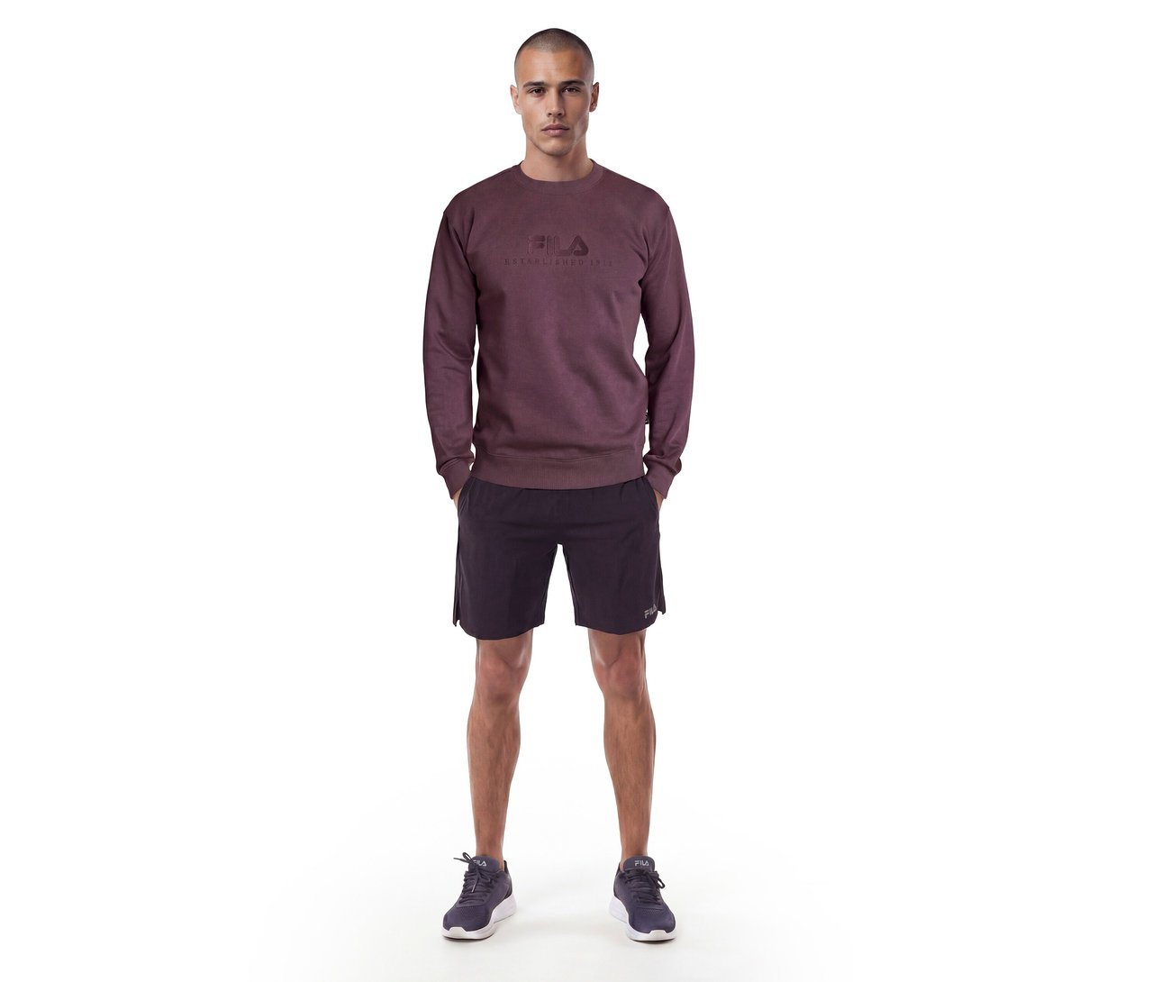Homme debout portant un sweat-shirt rouge foncé avec un logo Fila, un short noir et des baskets foncées.