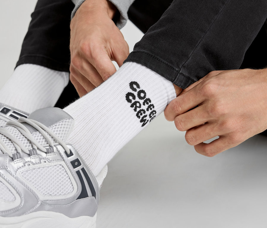 Un homme enfile des chaussettes blanches avec l'inscription « Coffee Crew » et des baskets argentées.