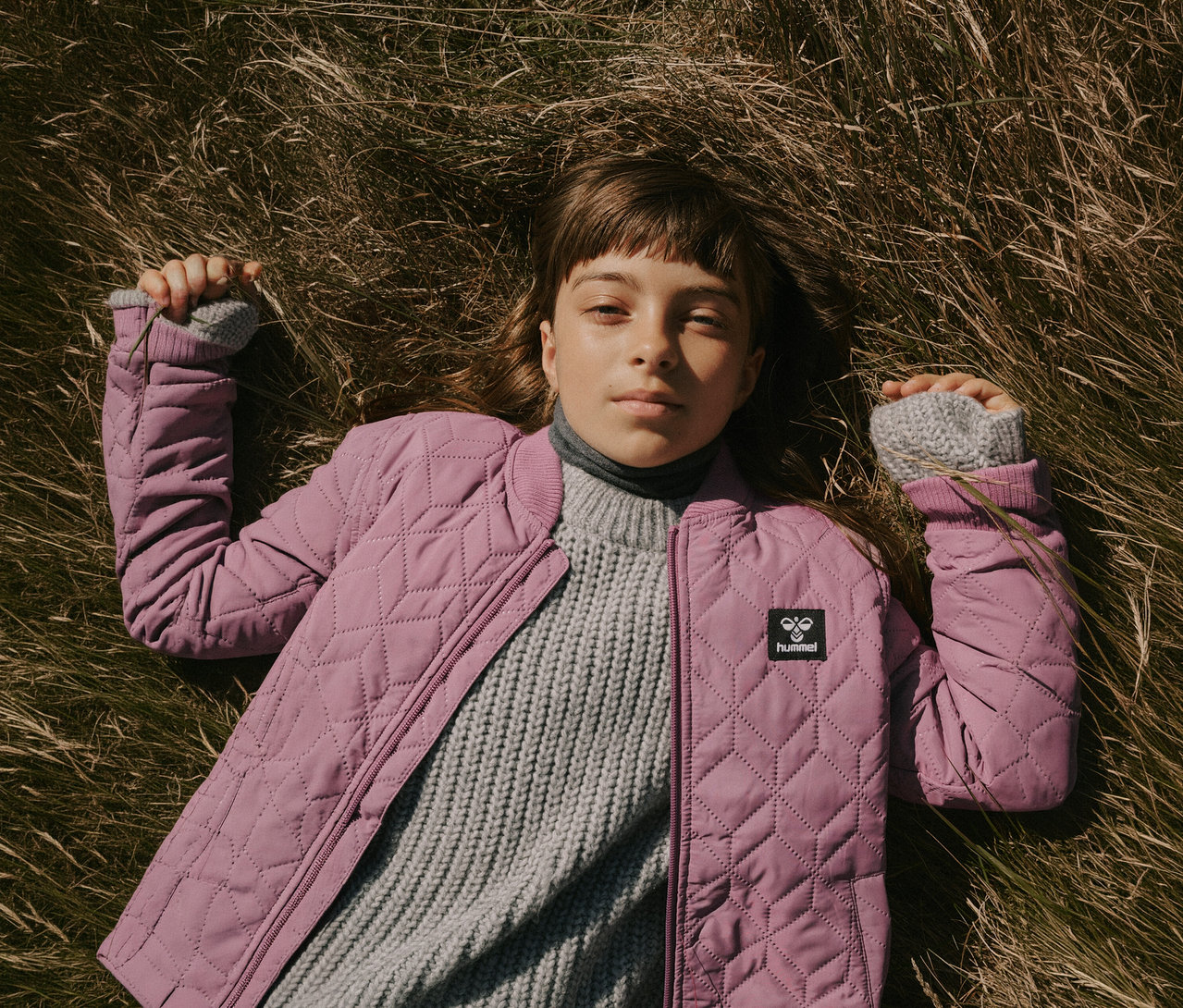 Une jeune personne est allongée sur le dos dans l'herbe sèche, portant une veste matelassée rose hummel® HMLSOBI Thermo-Set sur un pull gris, regardant l'appareil photo.