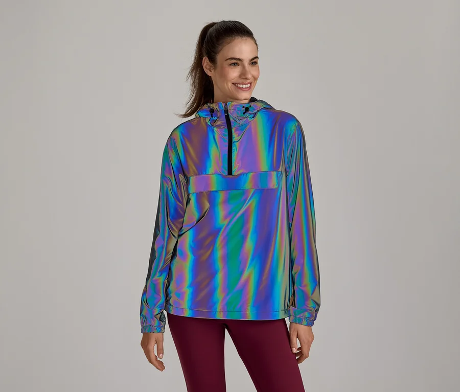 Femme souriante portant une veste anorak réfléchissante et irisée avec capuche et demi-zip, associée à un legging rouge foncé.