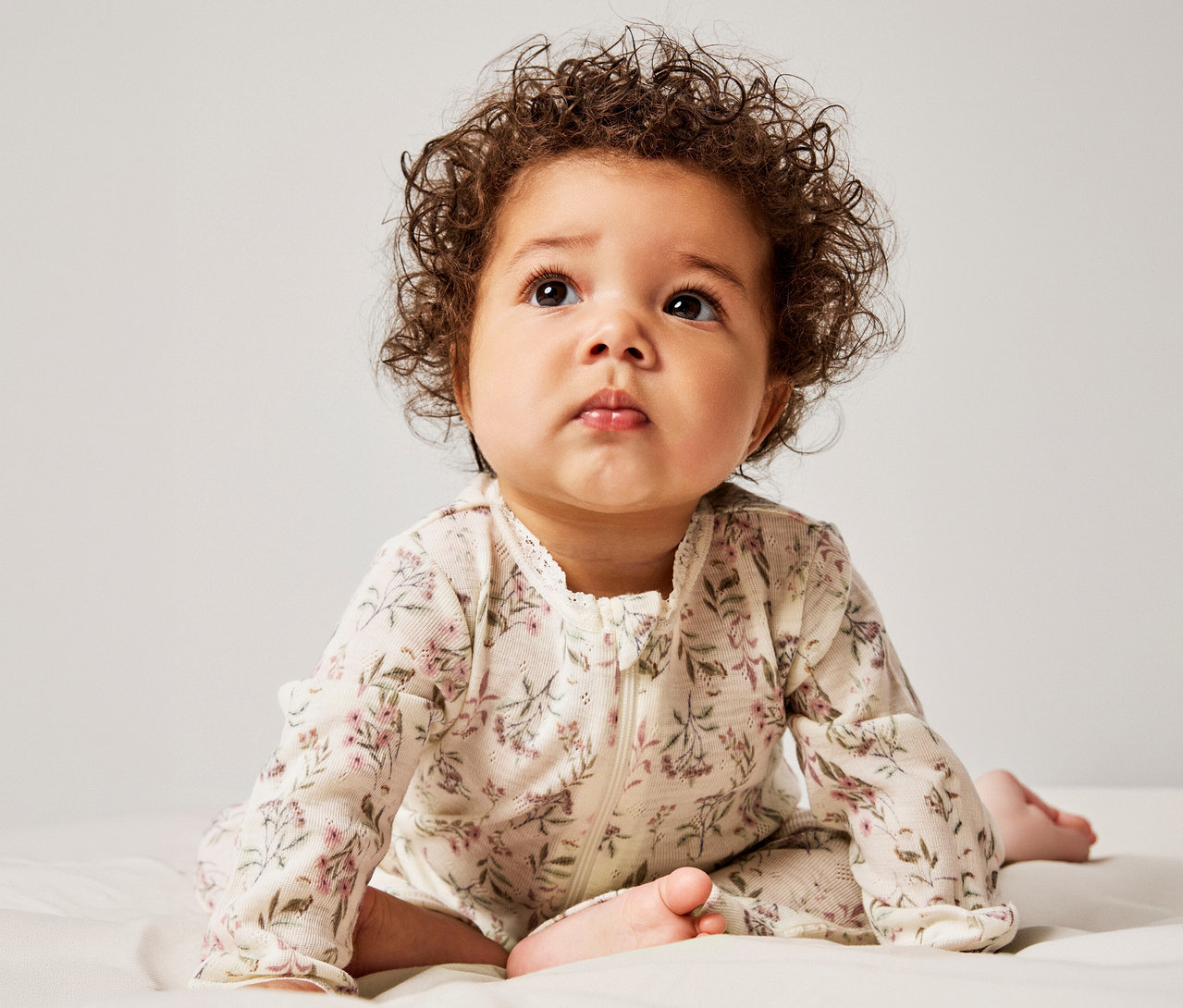 Un bébé aux cheveux bruns bouclés est allongé sur le ventre, regardant vers le haut. Il porte une grenouillère crème à manches longues avec un motif floral, une fermeture éclair et une bordure en dentelle.