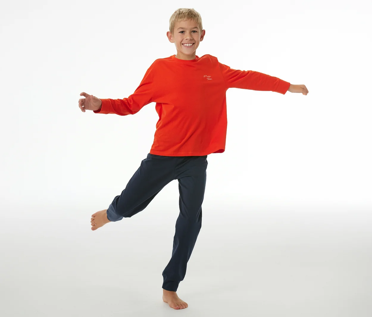 Garçon souriant portant un haut de pyjama enfant SCHIESSER rouge et un pantalon de pyjama bleu foncé avec des poignets, en équilibre sur une jambe, les bras tendus.