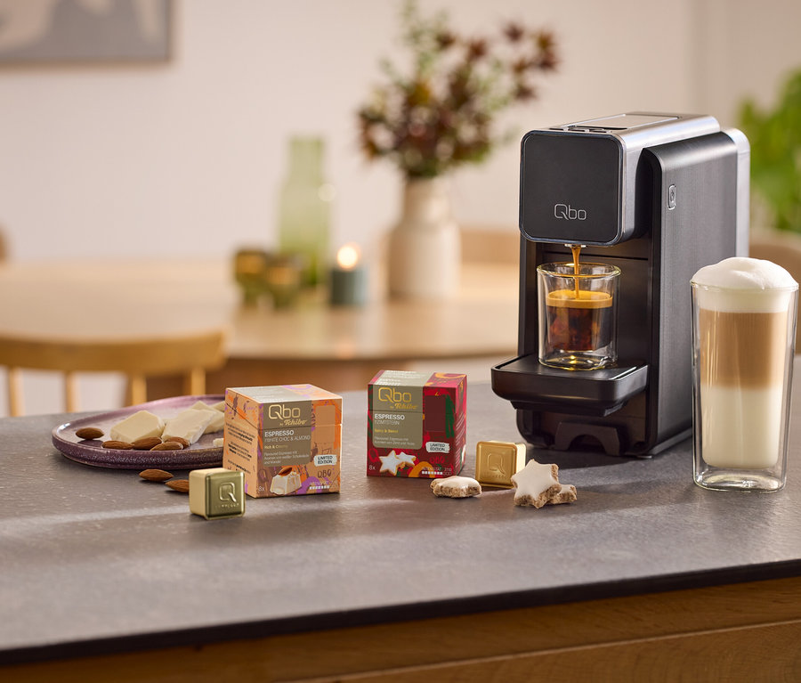 Une machine à capsules Qbo ONE noire prépare un espresso dans un verre, à côté d'un latte dans un grand verre. Sur le comptoir se trouvent des paquets Qbo Espresso White Choc Mandel, des capsules dorées, des biscuits étoiles, du chocolat blanc et des amandes.