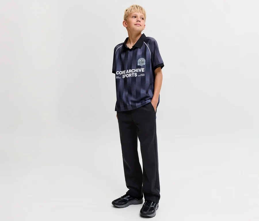 Un jeune garçon blond porte un polo rayé et un pantalon de survêtement noir Jack&Jones Bradley. Il se tient les mains dans les poches et regarde vers la droite.