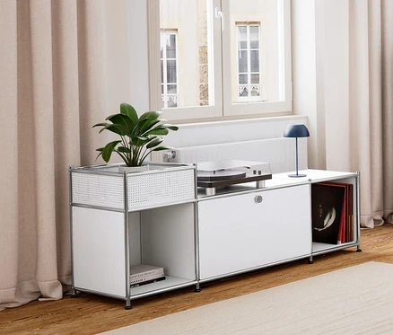Un salon avec parquet et tapis clair. À gauche, un meuble bas en métal « CN3 » blanc avec compartiment à abattant, surmonté d'une lampe de table à LED sans fil, bleu foncé. À côté, une commode en métal « CN3 » bleu foncé à 3 compartiments. Au centre, une table d’appoint avec plateau et fonction d’assise et un fauteuil. En arrière-plan, un buffet en métal « CN3 » gris clair à 3 abattants.