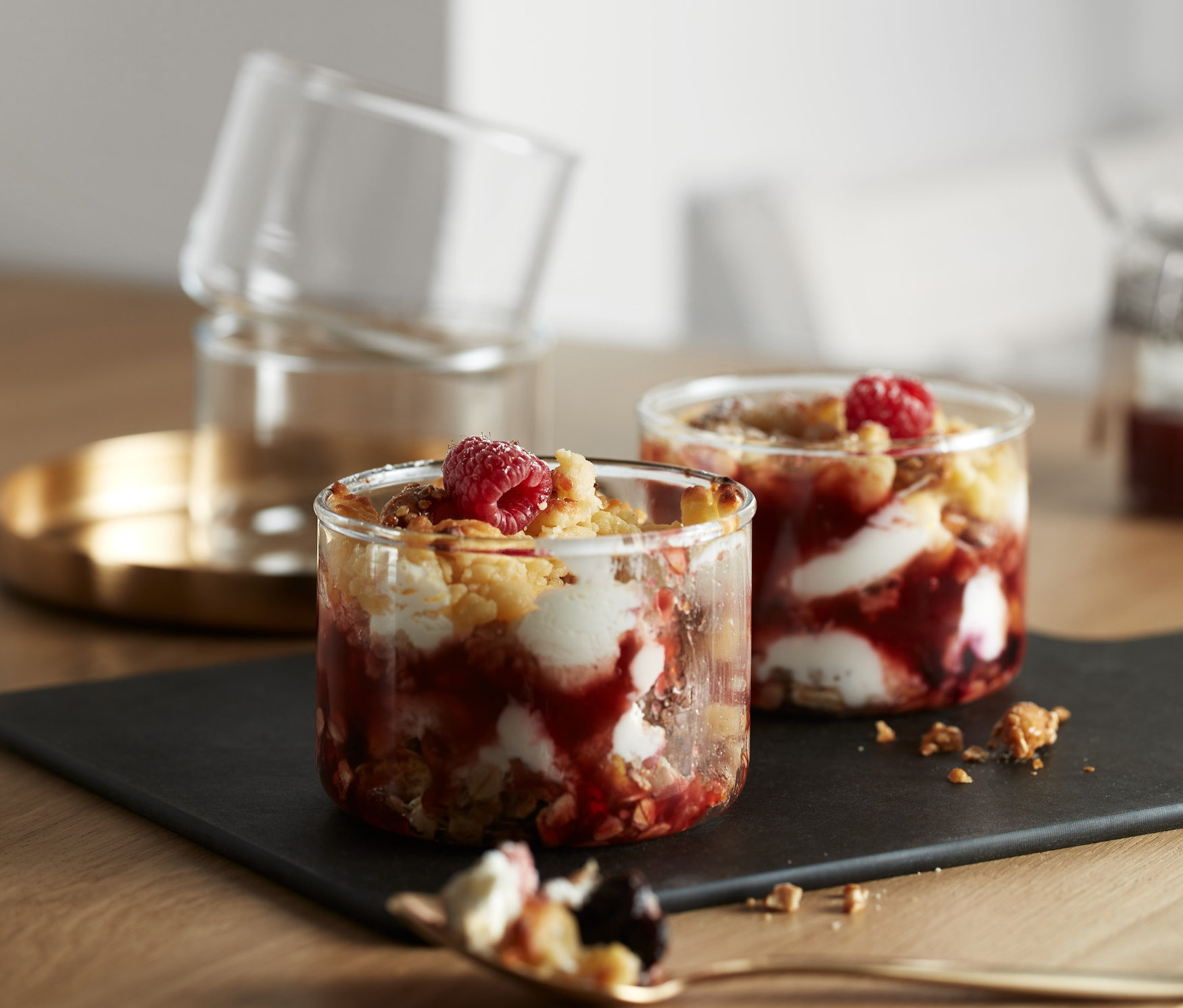 Sur une table, il y a 4 bocaux pour verrines ou préparations au four remplis d'un dessert aux fruits. Deux bocaux sont posés sur une ardoise noire, et deux autres sont empilés sur un plateau doré.