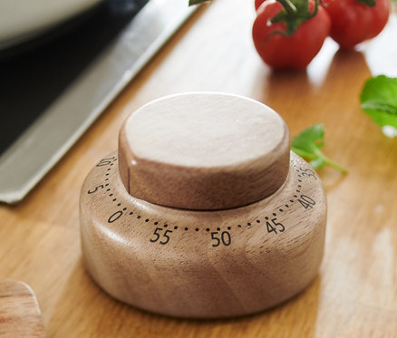 Sur une table en bois se trouvent un saladier, une minuterie en bois, des tomates cerises et une planche avec de la feta et des fleurs.