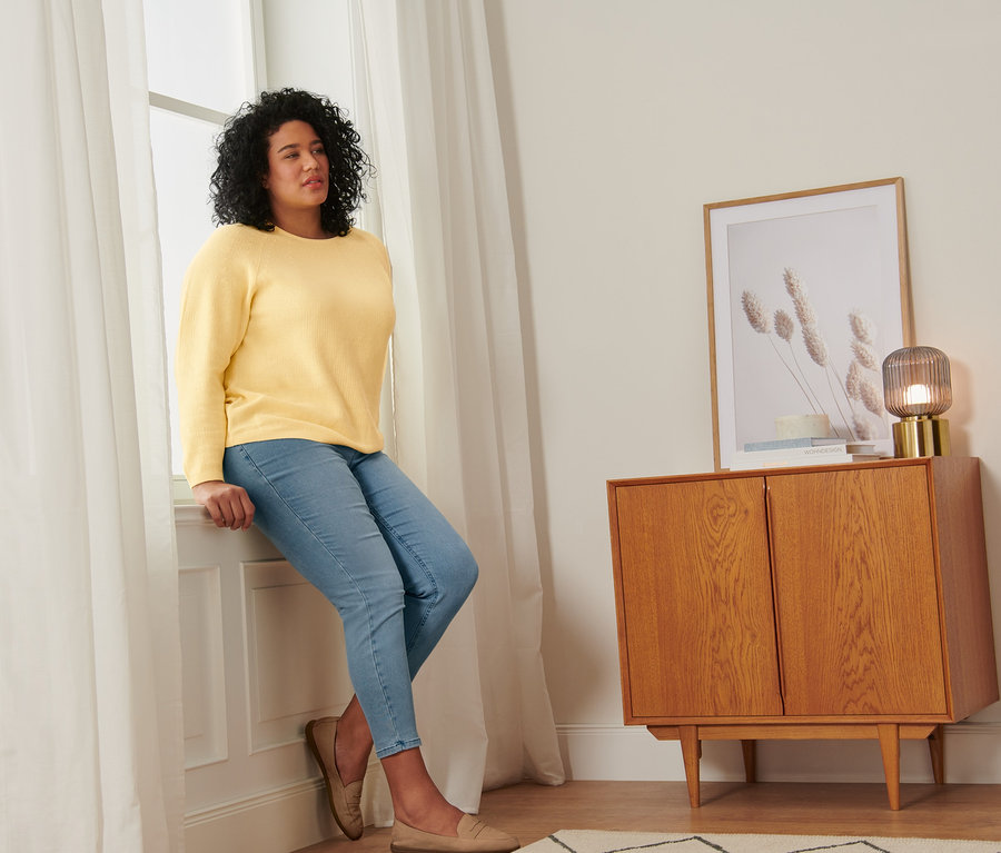 Une femme est assise sur un rebord de fenêtre portant un pull en maille jaune, un tregging et des mocassins en cuir velours.