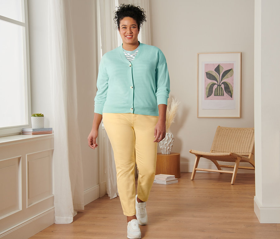 Une femme marche et porte un t-shirt à rayures vert menthe, un cardigan à encolure en V, un jean slim jaune et des baskets en cuir velours.