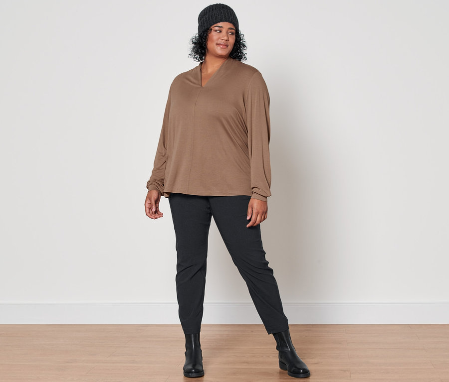 Une femme pose avec un t-shirt manches longues avec encolure en V beige, un pantalon prêt à porter en bengaline noir, des bottines en cuir et un bonnet en maille avec de la laine anthracite.