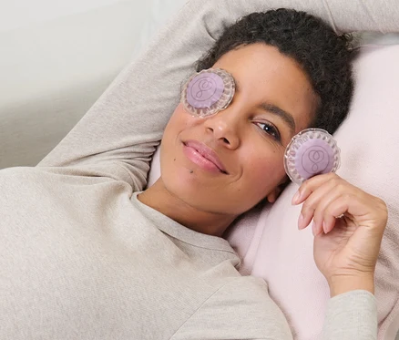 Femme allongée sur un canapé avec 2 tampons rafraîchissants pour les yeux, un sur l'œil et l'autre à la main.