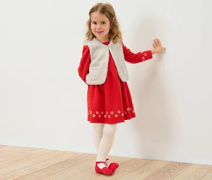 Une fille est appuyée contre un mur blanc et porte une robe rouge et un gilet sans manches enfant en polaire teddy.