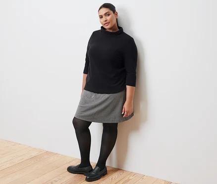 Une femme appuyée contre un mur porte un sweat-shirt manches 3/4 noir, une jupe molletonnée, des collants noirs et des loafers en cuir.