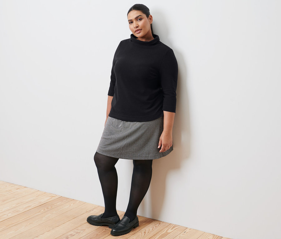 Une femme appuyée contre un mur porte un sweat-shirt manches 3/4 noir, une jupe molletonnée, des collants noirs et des loafers en cuir.