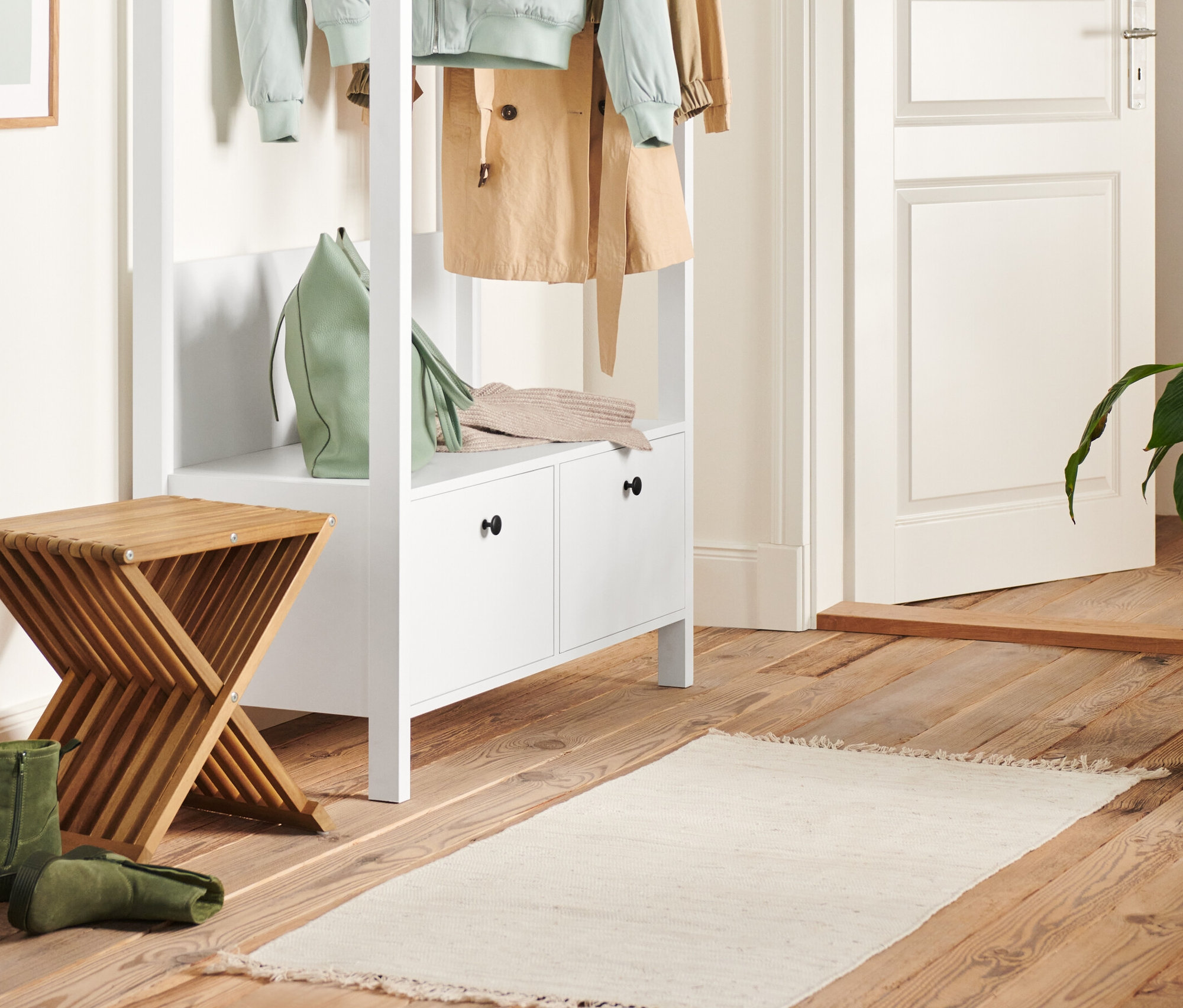 Dans le couloir, il y a une penderie blanche avec de grands tiroirs et un tabouret repliable. Un sac à main vert et un pull en tricot sont posés sur la penderie.