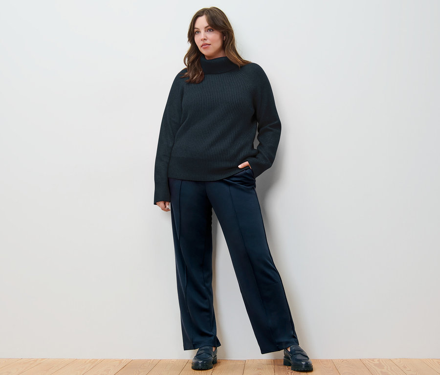 Une femme pose sur un plancher en bois portant un pull à col roulé en grosse maille bleu foncé et un pantalon en jersey avec passepoil.