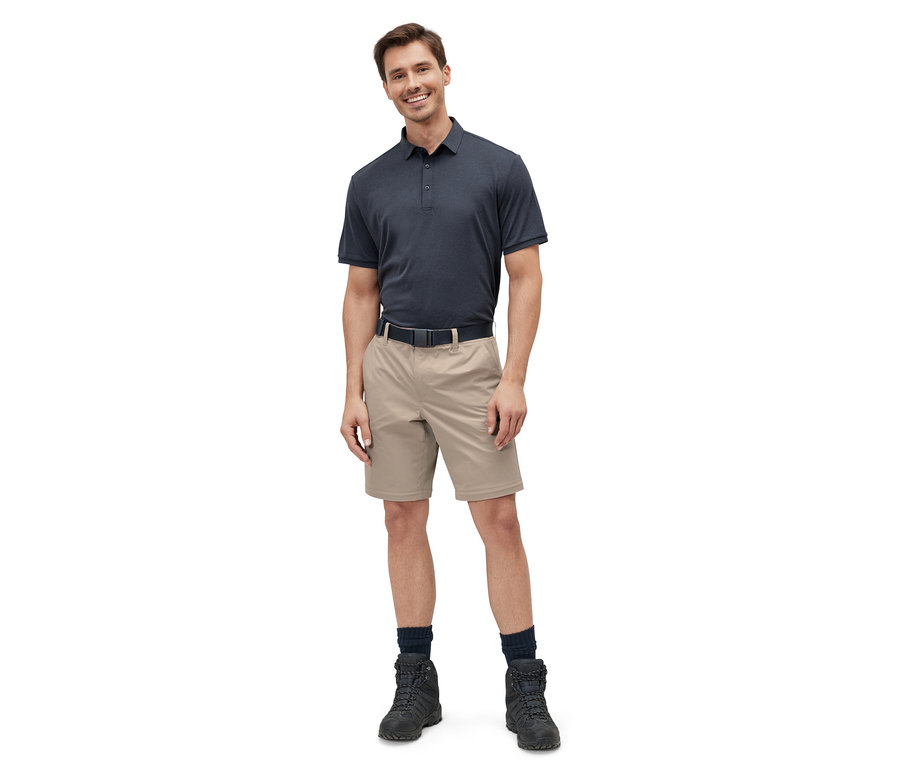 Un homme debout portant un pantalon technique 2 en 1 beige, un polo bleu marine, une ceinture noire et des bottes noires.