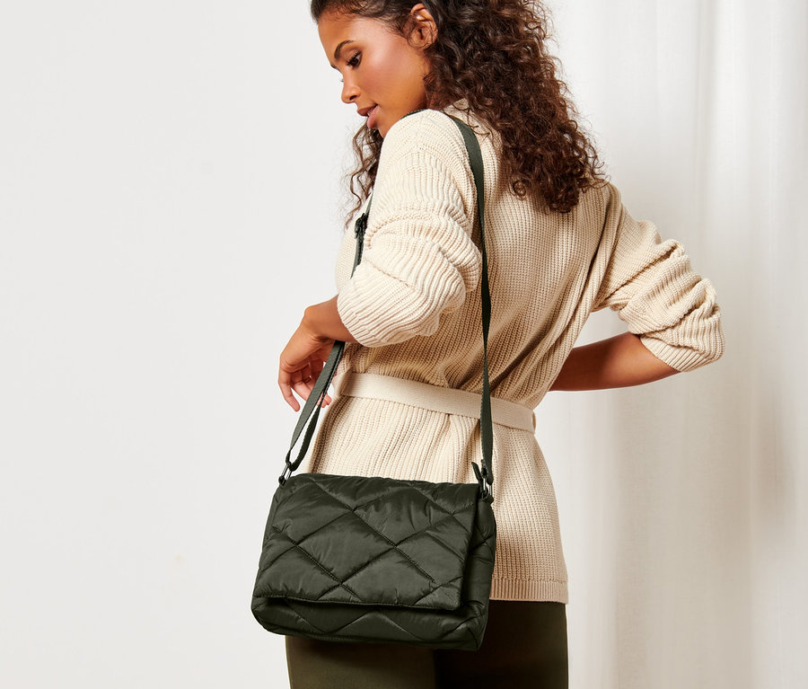 Une femme aux cheveux bruns bouclés portant un cardigan beige ceinturé pose avec un sac matelassé à bandoulière.