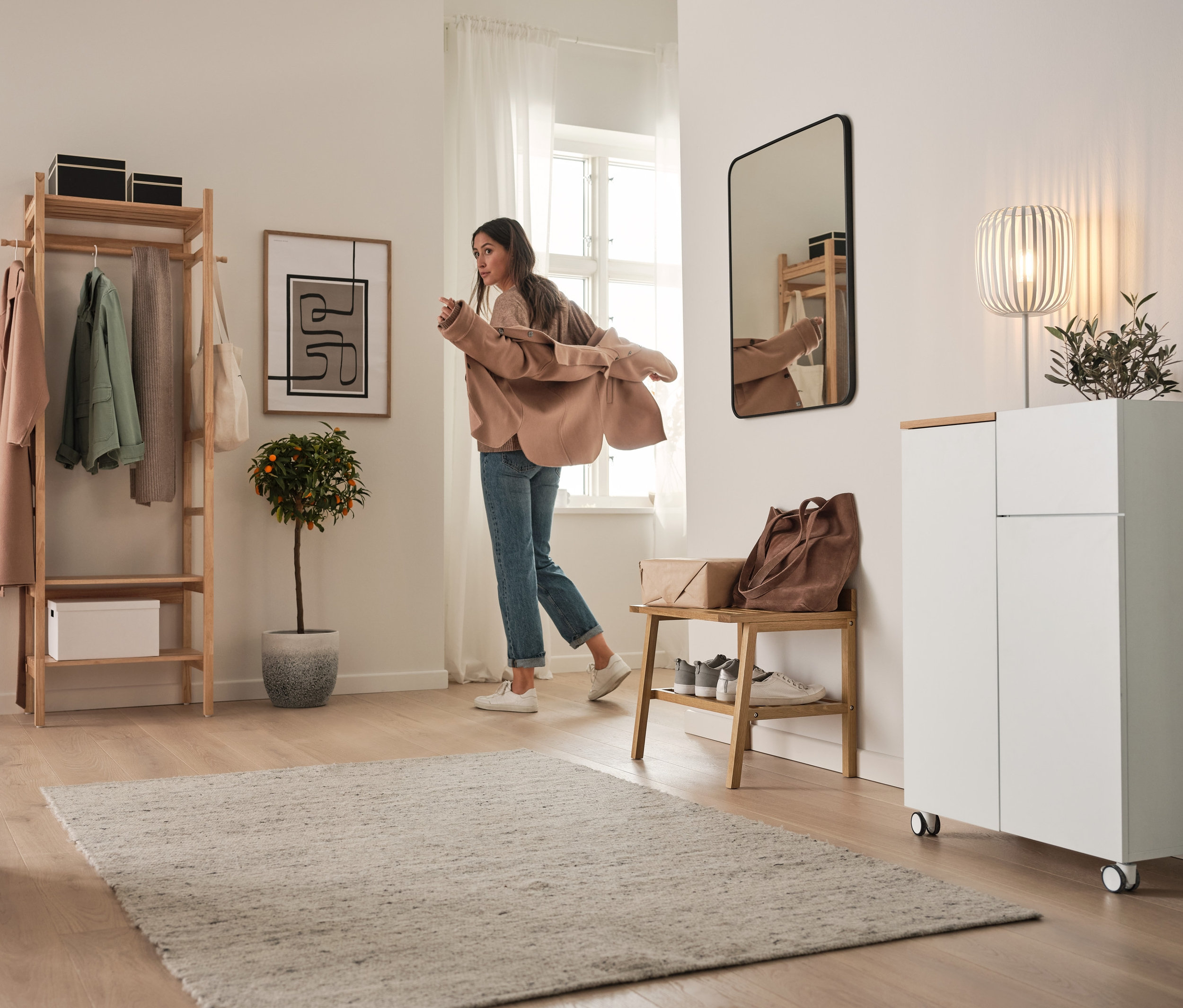Une femme enfile une veste dans un couloir. À l'arrière-plan, on peut voir un highboard à roulettes et une lampe de table en métal.