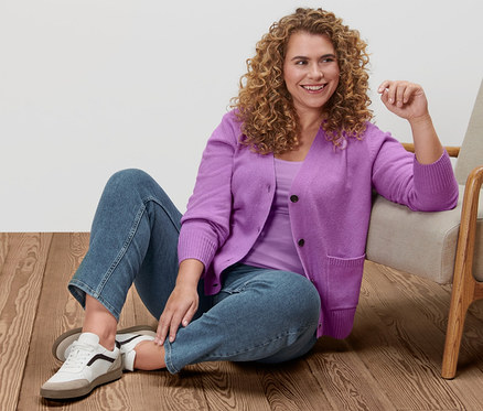 Une femme est assise par terre à côté d'un fauteuil et porte une jaquette en maille, un maillot de corps, un jean droit en denim et des baskets en cuir.