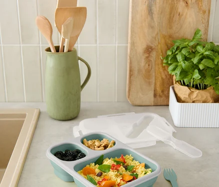 Une boîte à lunch bleue divisée avec des mûres, des noix et du couscous aux légumes est posée sur le comptoir.