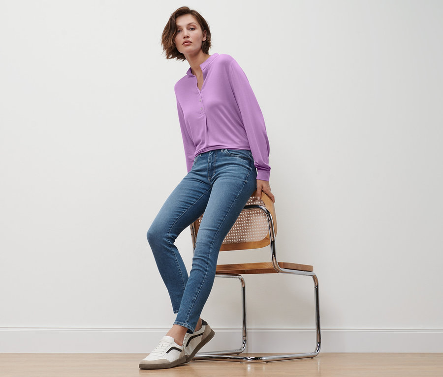 Une femme est assise sur une chaise et porte un jean slim en denim « Fit Emma », un chemisier lilas avec une patte de boutonnage et des baskets en cuir.