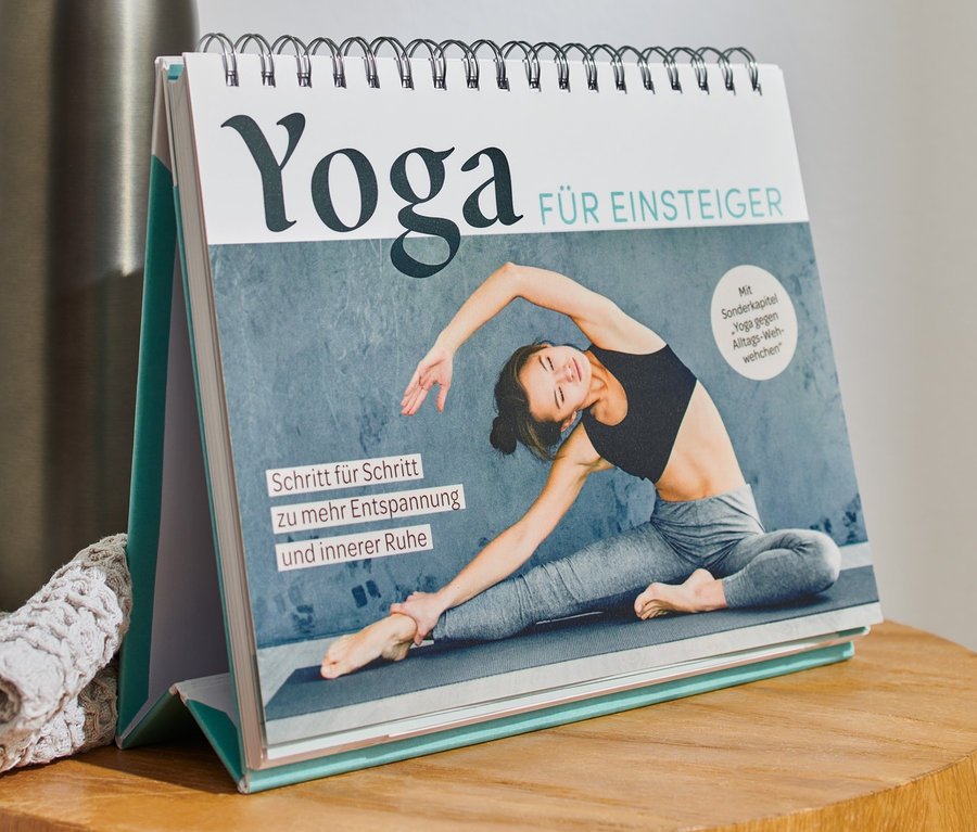 Sur une table en bois se trouvent un livre « Yoga für Einsteiger » (Le yoga pour les débutants, en allemand), une bouteille en acier inoxydable et un tissu tricoté gris.