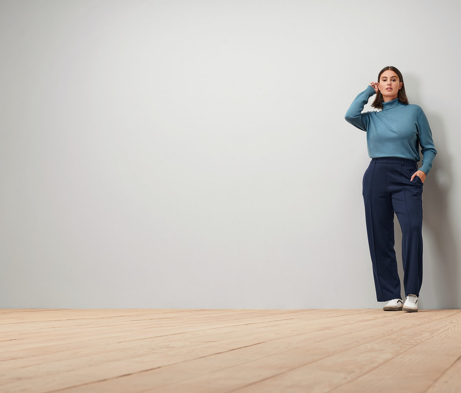 Femme posant en pull à col roulé en maille fine bleu fumée, pantalon en jersey bleu foncé avec nervure et baskets en cuir.