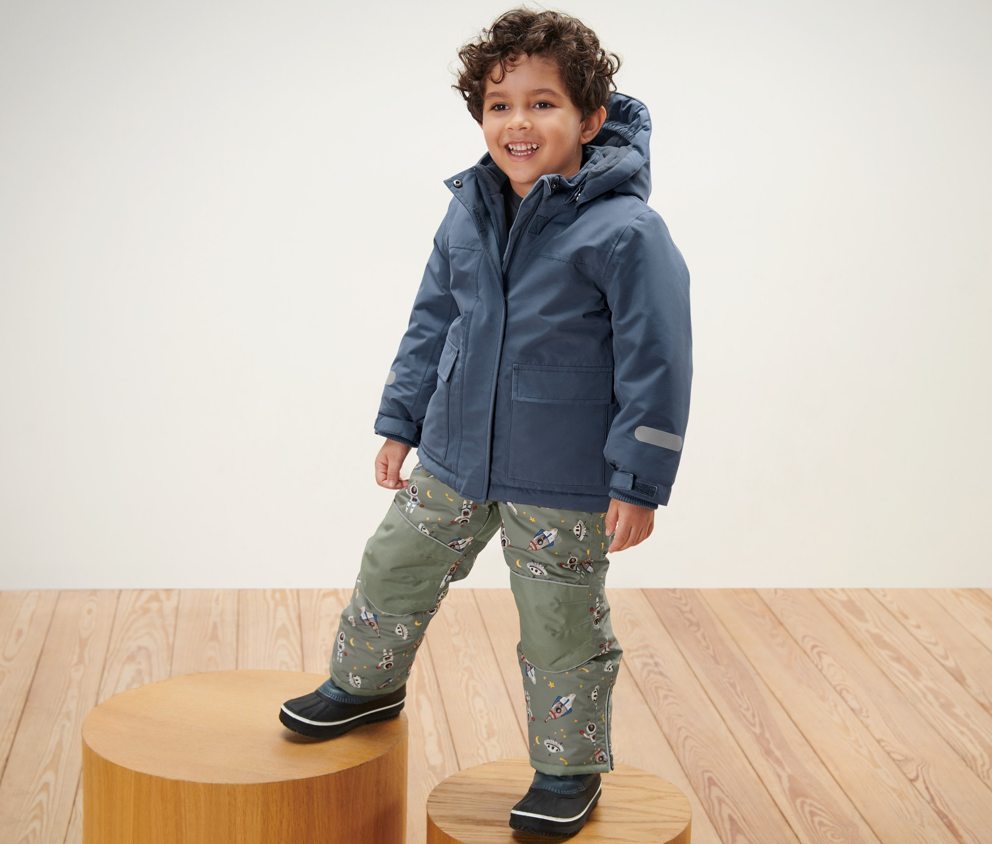 Un garçon debout sur un socle en bois porte une combinaison de ski pour enfant.