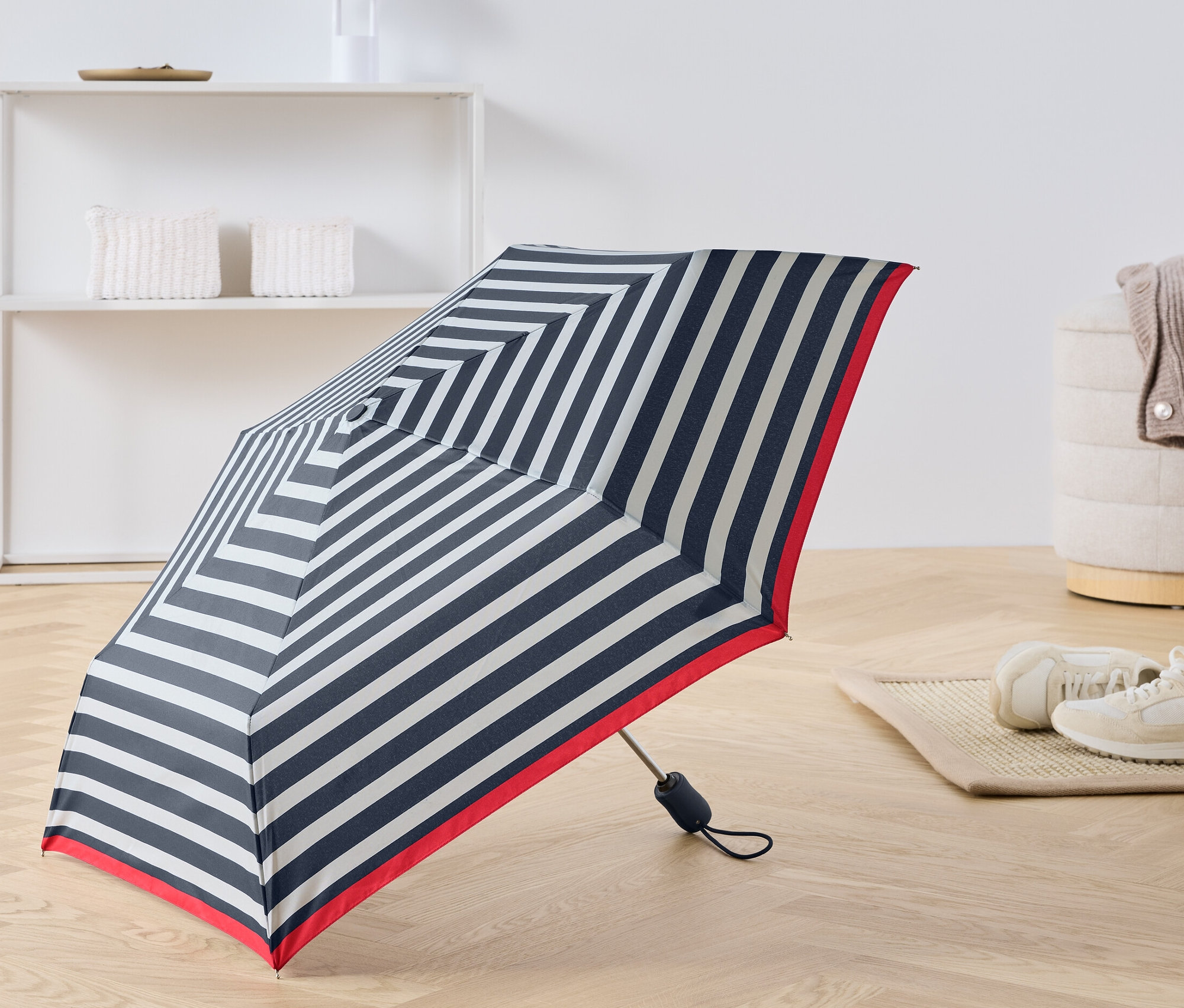 Un parapluie de poche automatique ouvert avec des rayures bleues et blanches et un bord rouge posé sur le sol.