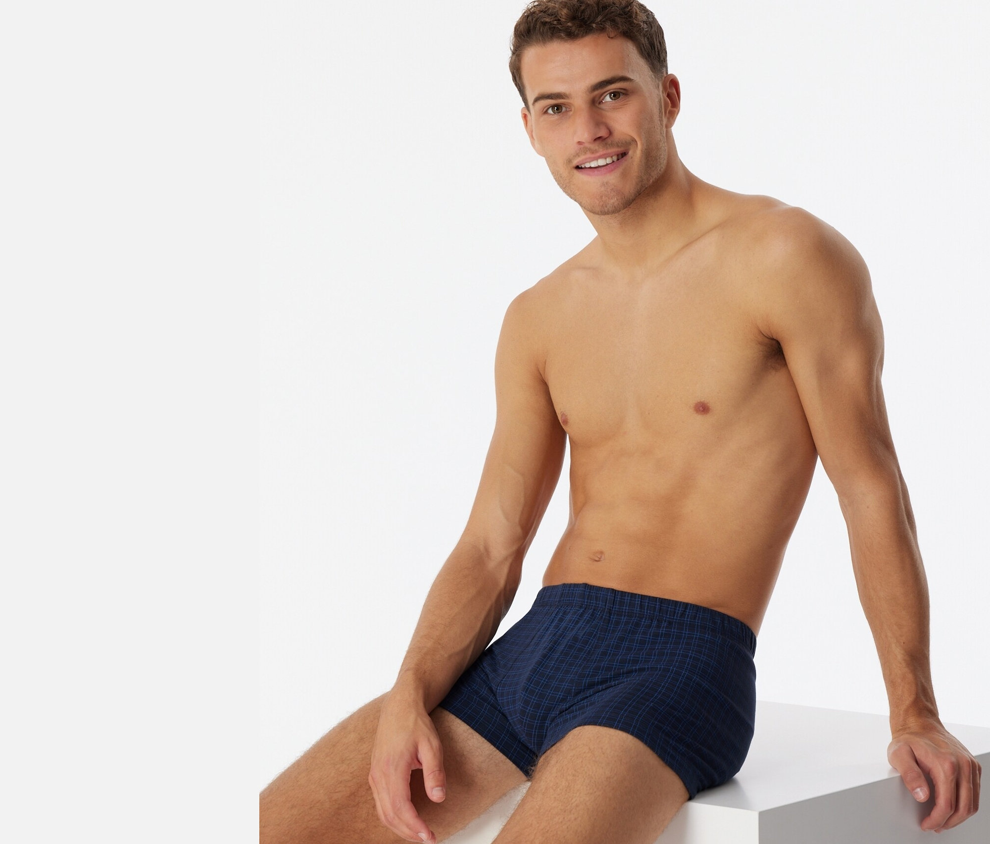 Un homme est assis sur un cube blanc et porte un short SCHIESSER à carreaux.