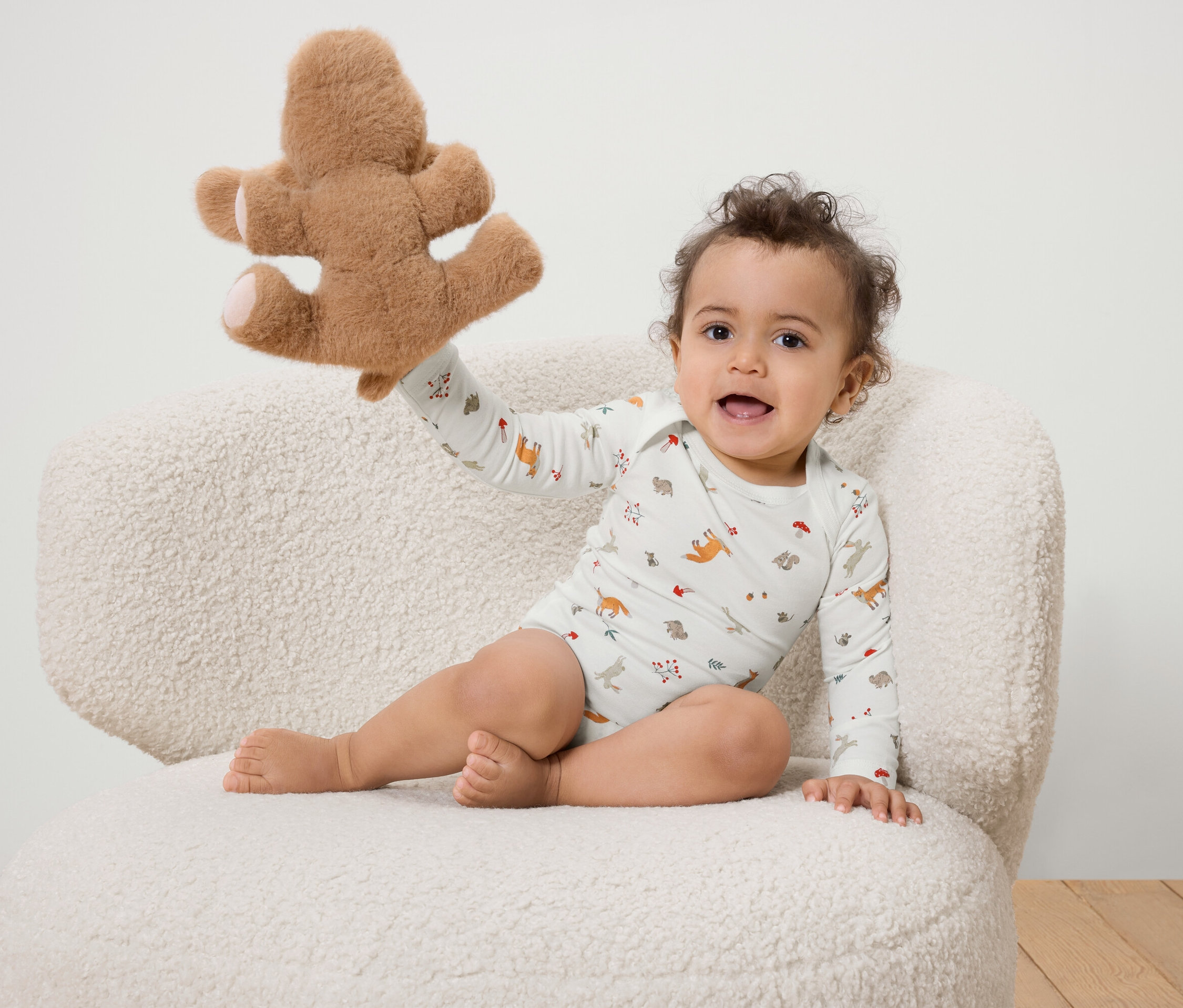 Un bébé souriant est assis sur une chaise blanche duveteuse et tient un ours en peluche.