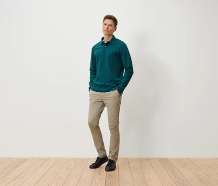 Un homme debout, les mains dans les poches, portant un polo vert à manches longues, un pantalon beige et des chaussures noires.