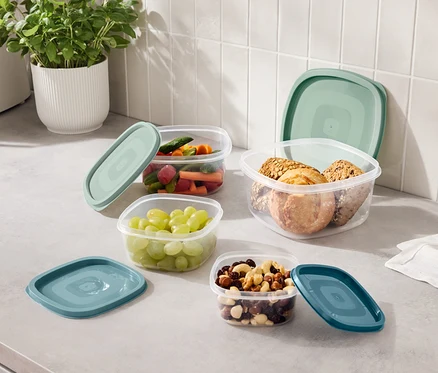 Quatre récipients transparents avec des couvercles bleu-vert remplis de raisins, de légumes coupés, de noix et de biscuits sont posés sur un comptoir de cuisine.