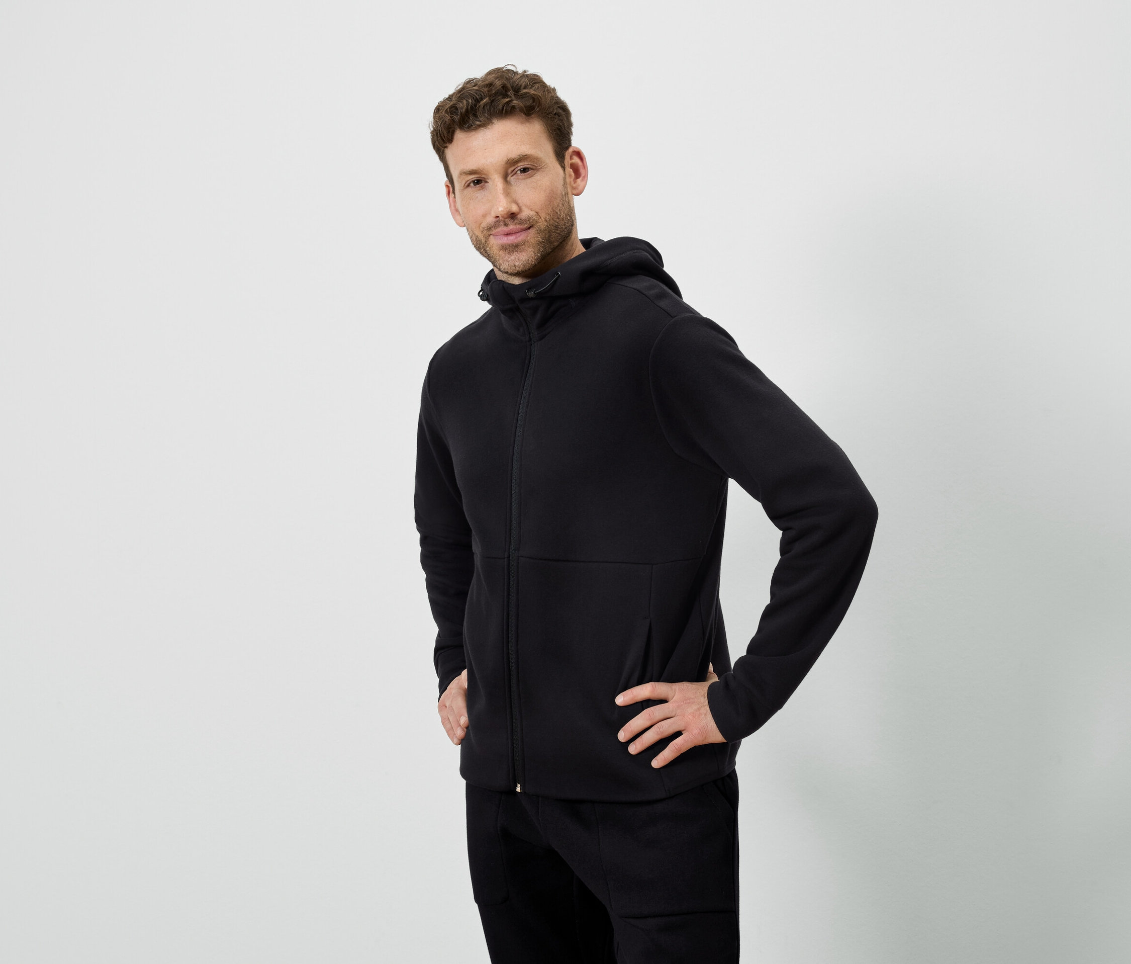 Un homme pose sur un fond blanc. Il porte un sweat à capuche noir et un pantalon noir.