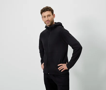 Un homme pose sur un fond blanc. Il porte un sweat à capuche noir et un pantalon noir.