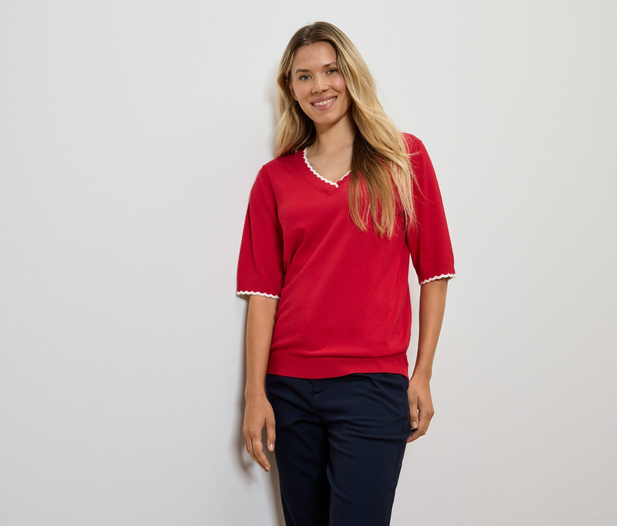 Une femme aux cheveux blonds pose en pull rouge et pantalon bleu marine.