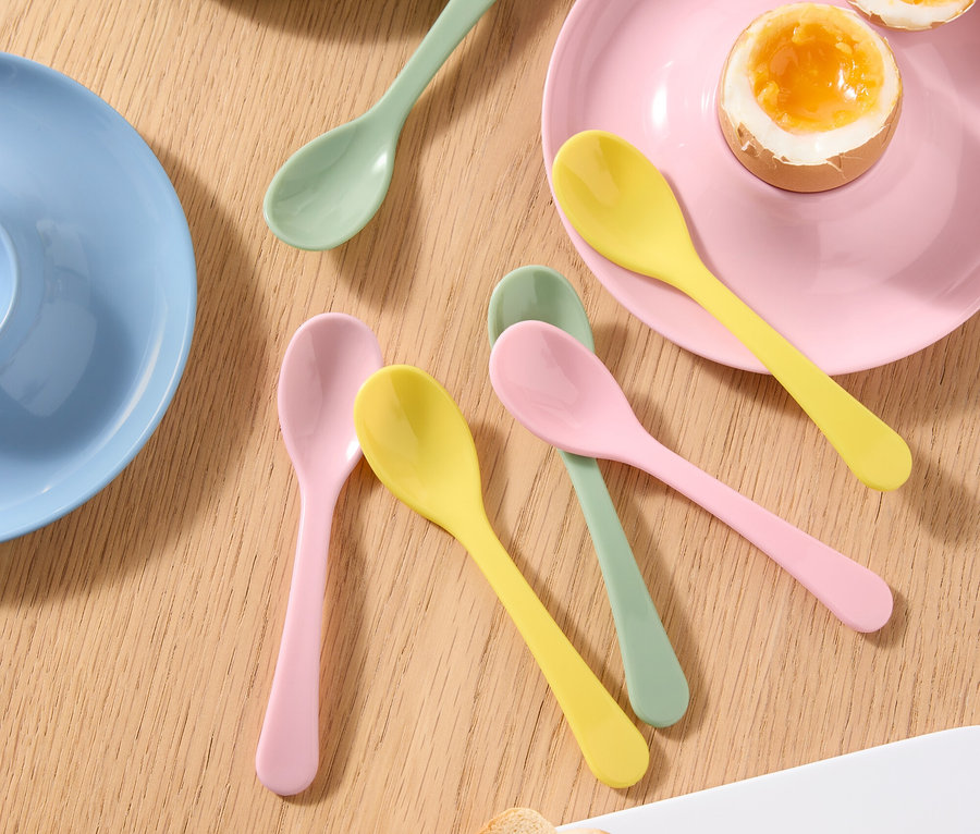 Vue de dessus d'une table avec un œuf à la coque dans un coquetier, un œuf à la coque coupé en deux dans une assiette, des cuillères colorées et des toasts.