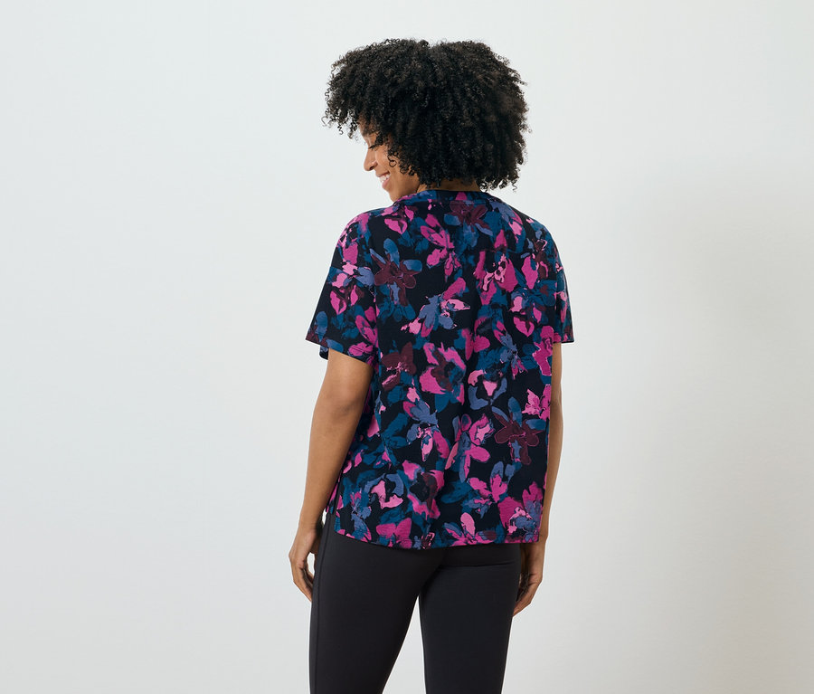 Une femme aux cheveux foncés, vue de dos, porte un chemisier noir à fleurs et un pantalon noir.