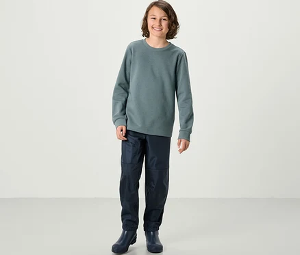 Un garçon debout portant un sweat-shirt bleu et un pantalon. 2 Kinder-Sweatshirts mit angerauter Innenseite, blau und salbei.