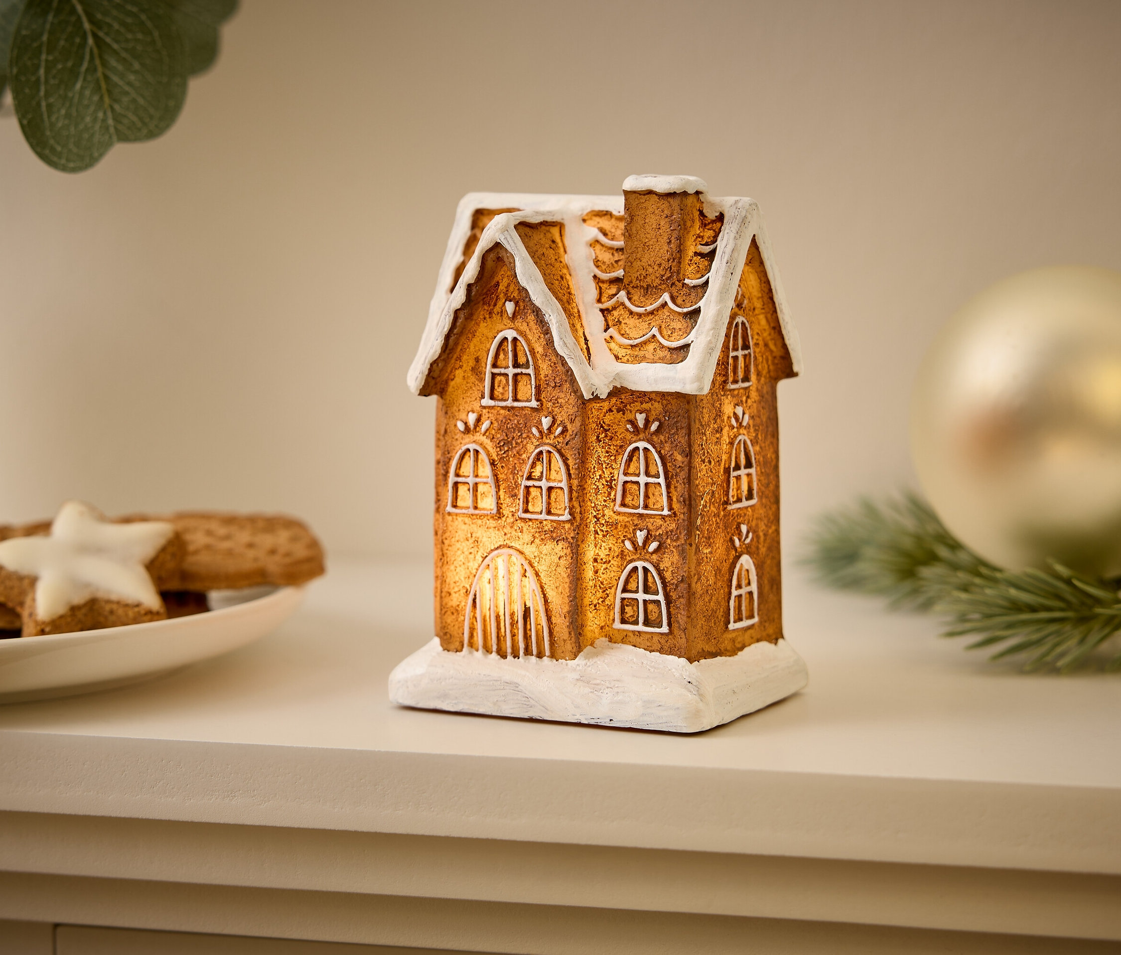 Une maison lumineuse décorative se dresse sur une étagère à côté de biscuits de Noël et d'une boule.