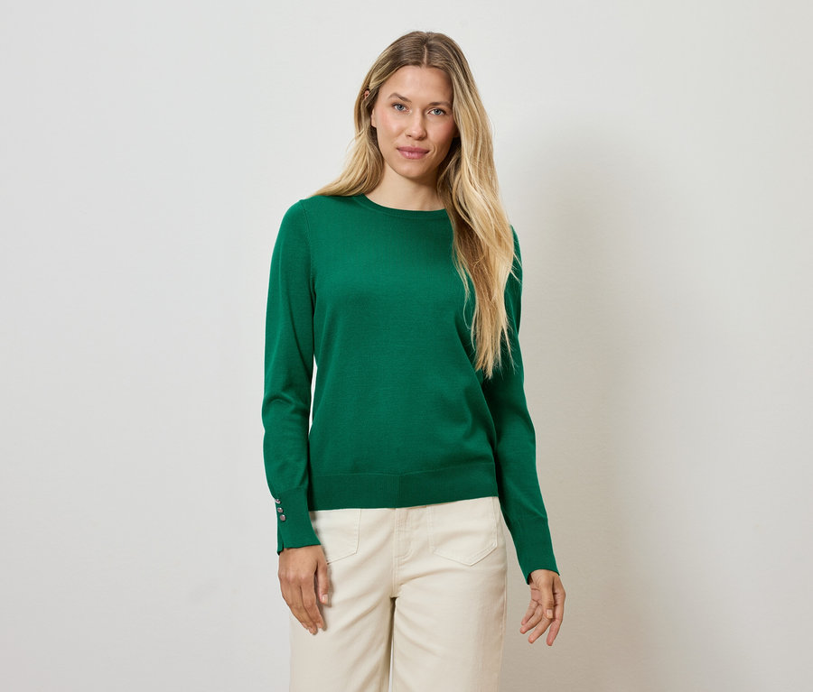 Une femme blonde portant un pull vert et un pantalon crème se tient devant un fond blanc.