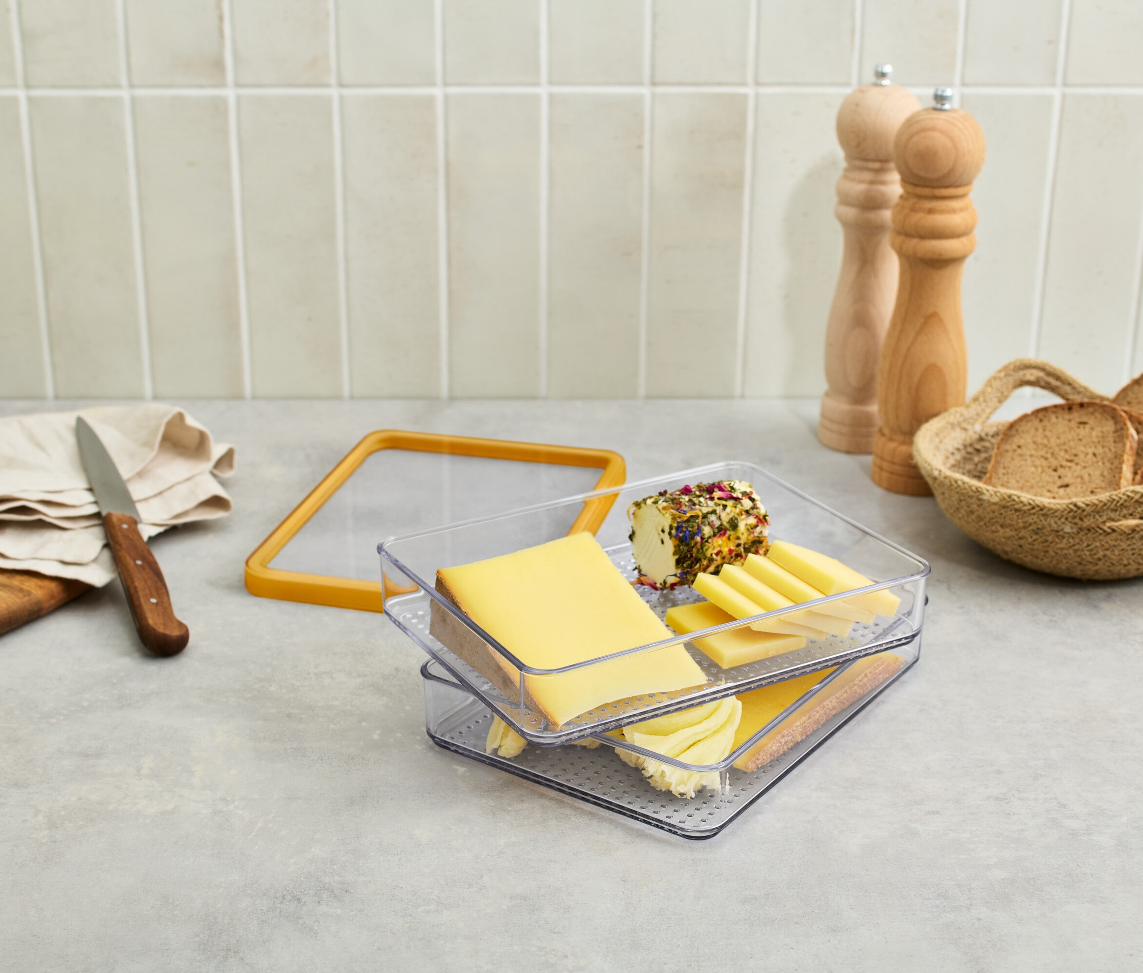 Sur une table, il y a du fromage dans un récipient transparent, des raisins, un couteau, du pain dans un panier et des moulins à épices.