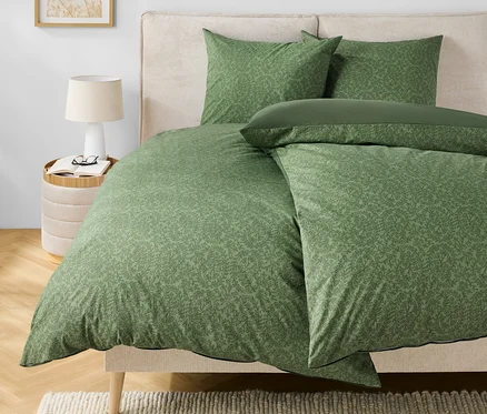 Linge de lit vert à motifs sur un lit avec une tête de lit beige. Une lampe et des lunettes sont posées sur la table de chevet.