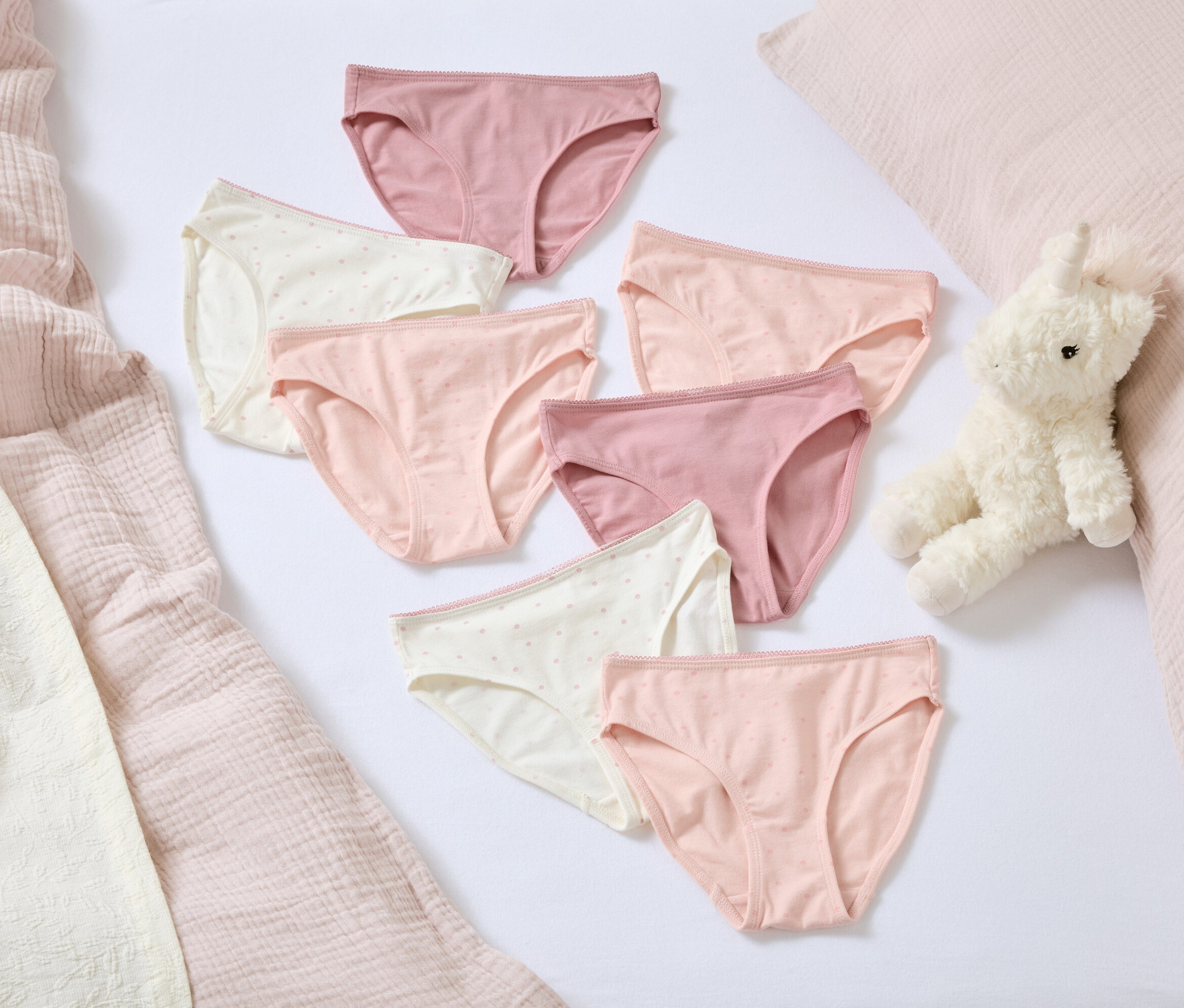 Sur un fond blanc, sept culottes de femmes dans différentes nuances de rose et de blanc. À côté d'eux se trouvent un lapin en peluche et un oreiller rose.